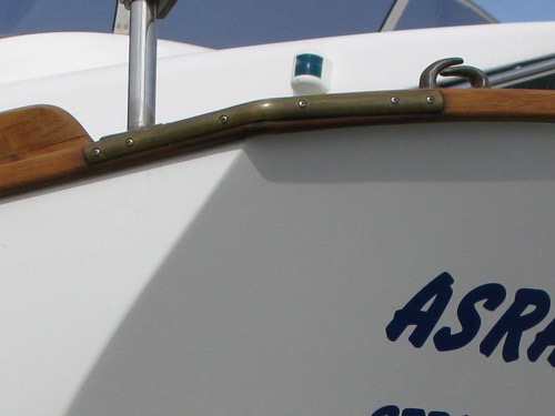 BIANCA 27's flotte lønningsliste af massiv teak hele skibet rundt er en markant del af bådens profil. Forrest er listen beskyttet af stævnbeslaget i rustfrit stål. Agterude ses her en flot løsning på at beskytte lønningen, et elegant udformet messingbeslag, der dækker hele hjørnet og matcher fint til klydset af messing. Set på DEN-80 Asra fra Struer. 